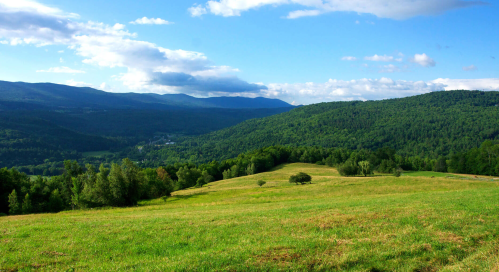 A scenic landscape featuring rolling green hills, mountains in the distance, and a bright blue sky with clouds.