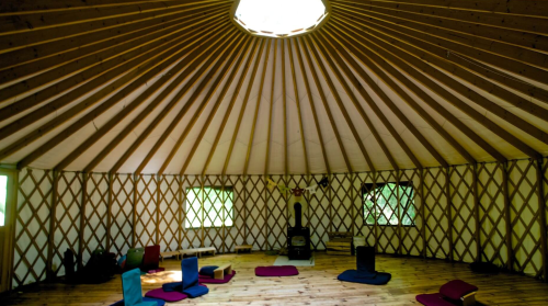 Interior of a yurt with wooden beams, a circular skylight, and colorful cushions arranged on a wooden floor.