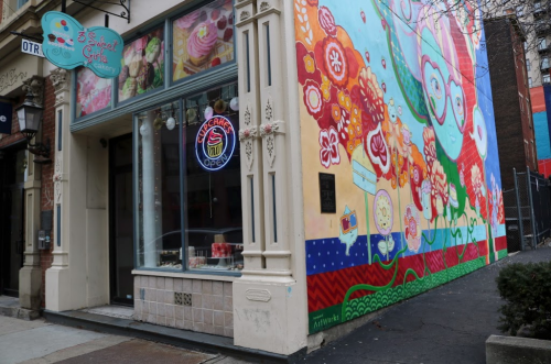 Colorful mural on a building exterior with a bakery entrance featuring a neon "Open" sign.