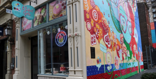 Colorful mural on a building next to a dessert shop with a sign that says "Sweet Treats" and an open cupcake display.