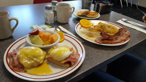 A breakfast table with eggs Benedict, fruit cup, and a plate of bacon and pancakes. Coffee mugs in the background.
