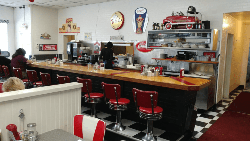 A retro diner interior with red and white decor, a long counter, and vintage Coca-Cola signage.