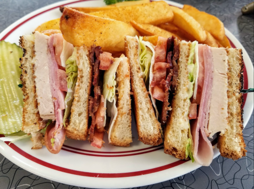 A plate with a stacked sandwich featuring layers of turkey, ham, lettuce, and tomato, alongside crispy potato wedges.