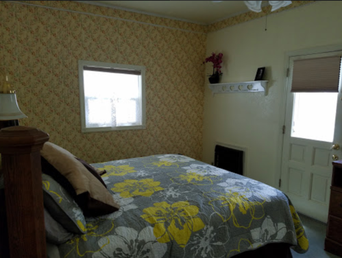 Cozy bedroom with floral bedding, a window, and a door, featuring light-colored walls and a decorative shelf.