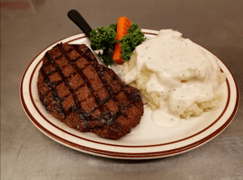 A grilled steak with a knife, served alongside mashed potatoes with gravy and a side of kale and carrot.