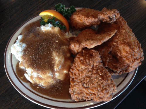 A plate of fried chicken with mashed potatoes and gravy, garnished with a slice of orange and a sprig of kale.
