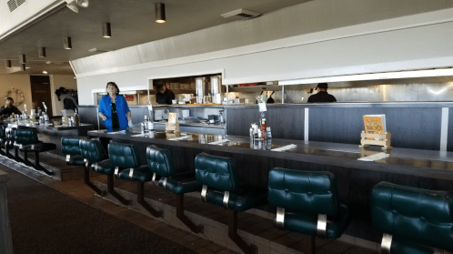 A diner interior featuring a long counter with green stools and a staff member serving behind the counter.