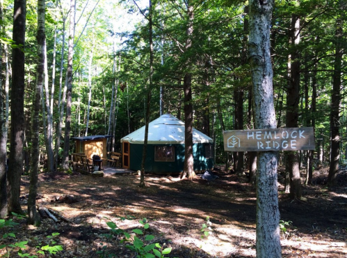 A green yurt nestled in a forest, with a wooden sign reading "Hemlock Ridge" nearby.