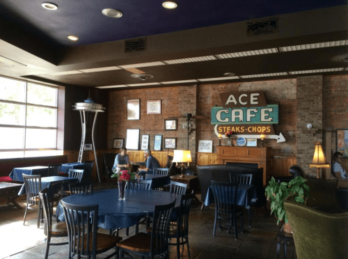 Interior of Ace Cafe with tables, chairs, and a brick wall featuring a colorful sign. Warm lighting and decor create a cozy atmosphere.
