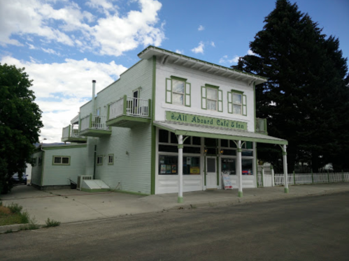 A two-story white building with green accents, featuring a café and inn sign, surrounded by trees and a blue sky.