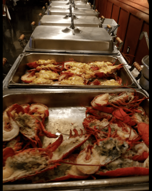 A buffet display featuring trays of cooked lobster and seafood, with some topped with cheese.
