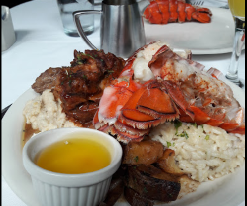 A plate of seafood and meat featuring lobster, grilled meat, rice, and potatoes, with a small cup of sauce on the side.