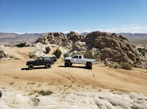 Two off-road vehicles parked on a sandy trail near rocky terrain under a clear blue sky.