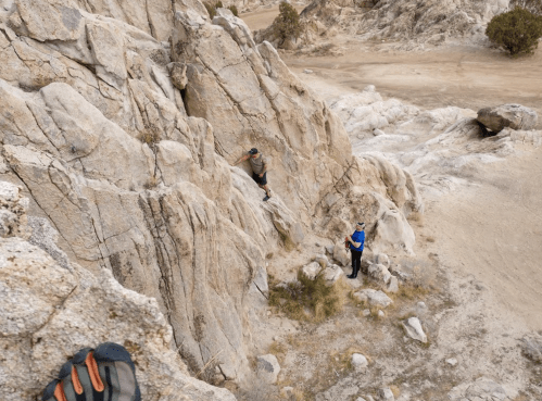 Two climbers navigate rocky terrain in a desert landscape, with one scaling a cliff and the other observing below.