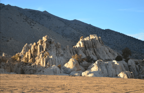 Rocky formations under a clear sky, with mountains in the background and a sandy foreground.