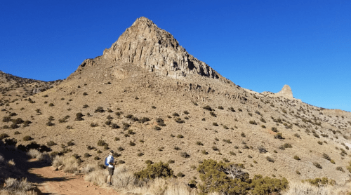 A hiker stands on a trail in a desert landscape, with a prominent rocky peak and clear blue sky in the background.