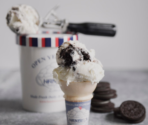 A scoop of ice cream in a cone beside a tub of Handel's ice cream and Oreo cookies on a marble surface.