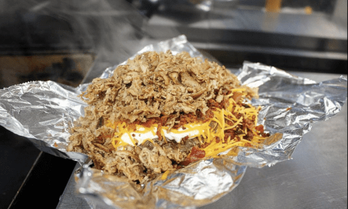 A foil-wrapped dish piled high with crispy fried meat, cheese, and toppings, emitting steam.
