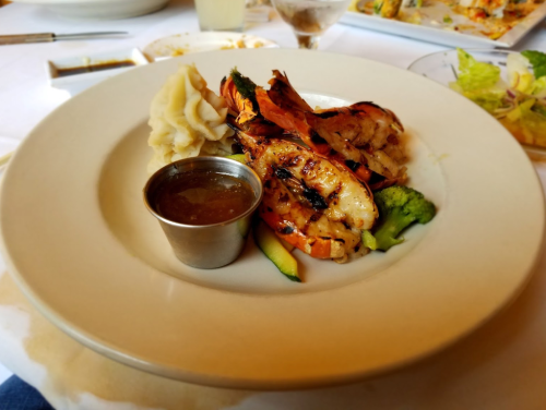 Grilled shrimp on a plate with mashed potatoes, broccoli, and a small cup of sauce.