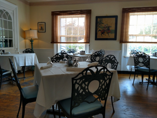 A cozy dining room with white tablecloths, decorative chairs, and large windows letting in natural light.