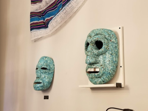 Two mosaic masks in shades of blue displayed on a wall, with a colorful textile hanging above them.