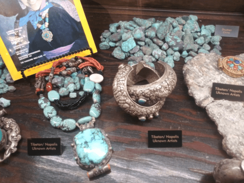 Display of turquoise jewelry and stones, featuring necklaces, bracelets, and a photo of Tibetan/Navajo artists.