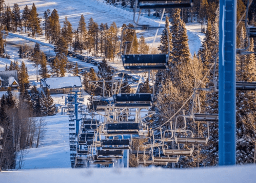 A snowy ski resort scene featuring empty chairlifts and pine trees under a clear blue sky.