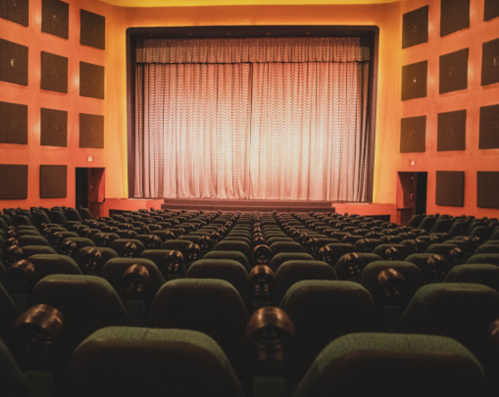 Interior of a theater with green seats facing a stage, featuring a closed curtain and warm orange walls.