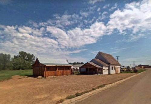 A rural scene featuring two wooden buildings under a blue sky with scattered clouds, alongside a dirt road.
