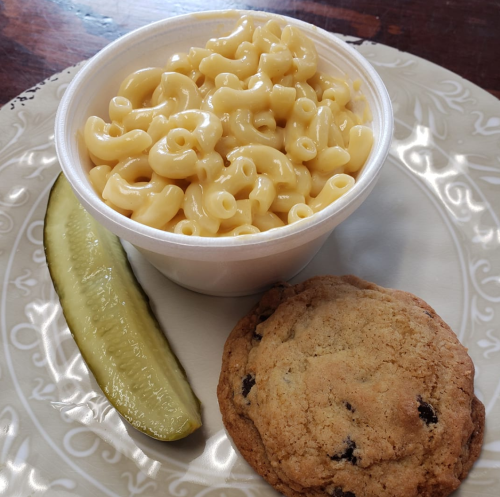 A plate with a cup of macaroni and cheese, a pickle slice, and a chocolate chip cookie.