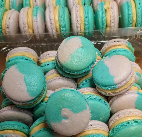 A close-up of colorful macarons in shades of teal, gray, and yellow, stacked in a clear container.