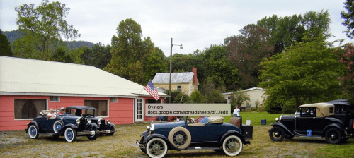 A vintage car gathering in a rural setting with a pink building and trees in the background.
