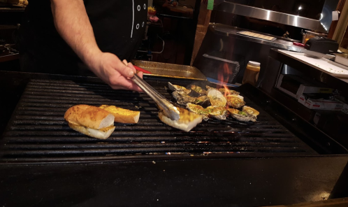 A person using tongs to grill bread and oysters on a barbecue, with flames visible in the background.