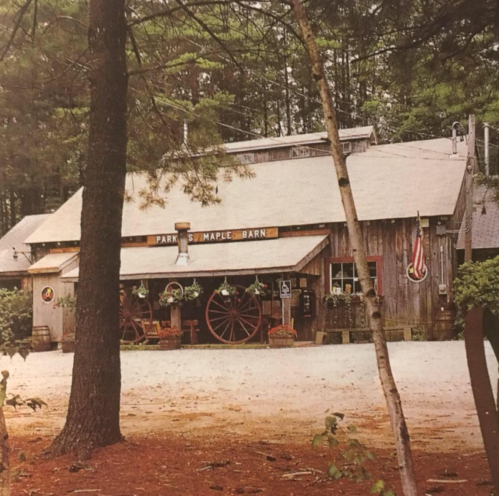 A rustic wooden barn surrounded by trees, featuring a sign that reads "Parker's Maple Barn" and decorative planters.