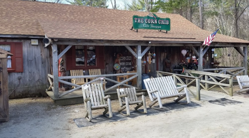 A rustic gift shop with a wooden exterior, rocking chairs, and an American flag outside, surrounded by trees.