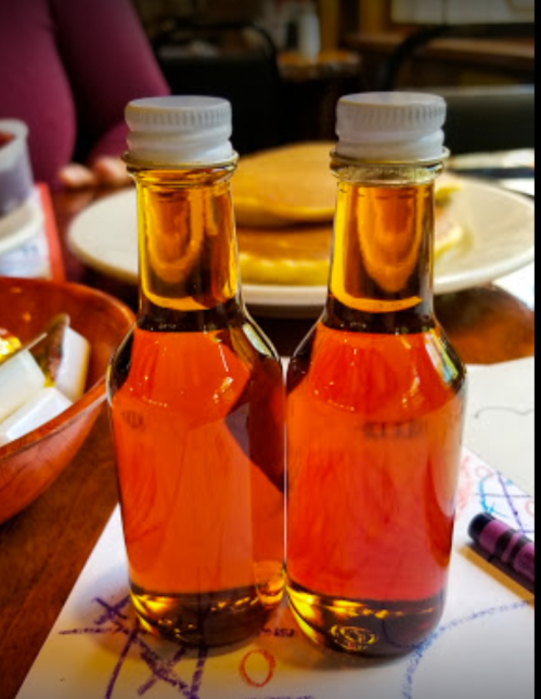 Two small glass bottles of syrup sit on a wooden table, with pancakes visible in the background.
