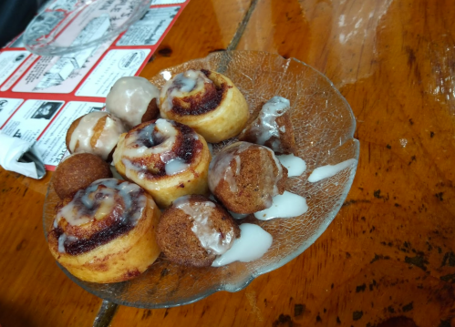 A plate of cinnamon rolls and small doughnut holes drizzled with white icing on a wooden table.
