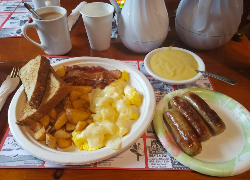 A breakfast plate with scrambled eggs, bacon, potatoes, toast, and sausages, alongside coffee in cups.