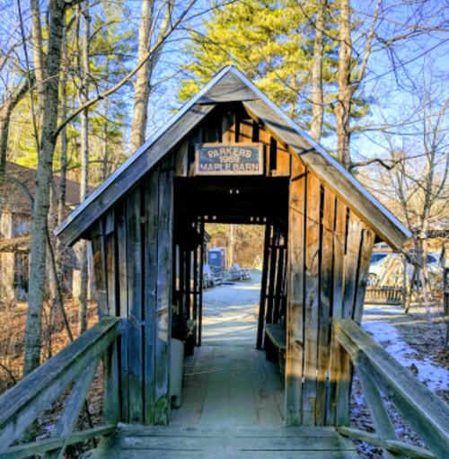 A wooden bridge leads to a rustic barn entrance surrounded by trees in a serene outdoor setting.