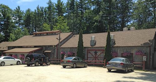 A rustic wooden building with a gravel parking lot, surrounded by trees and flower planters, featuring two parked cars.