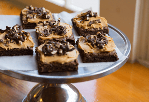 A silver cake stand holds brownies topped with creamy peanut butter frosting and crushed nuts.