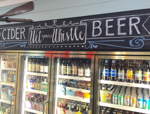 Chalkboard sign above a beverage fridge reads "CIDER Let your Whistle BEER," with various drinks visible inside.