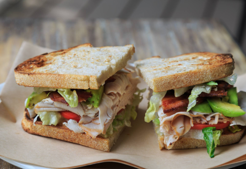 A close-up of a toasted sandwich filled with turkey, lettuce, tomato, and avocado, served on a paper plate.