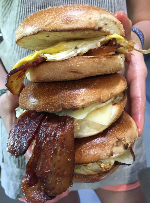 A person holds a towering stack of bagels filled with bacon, cheese, and an egg, showcasing a hearty breakfast.