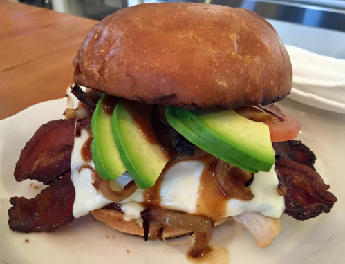 A close-up of a delicious sandwich with bacon, cheese, avocado, tomato, and grilled onions on a toasted bun.