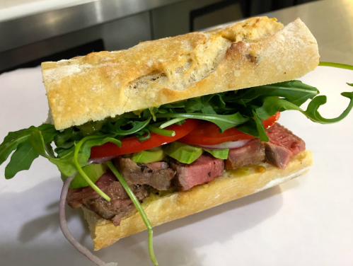 A fresh sandwich with layers of sliced beef, tomatoes, arugula, and green onions on a crusty bread roll.