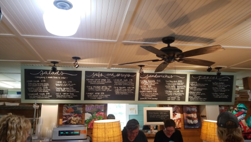 Menu boards displaying salads, wraps, sandwiches, and paninis in a cozy café setting with patrons in the background.