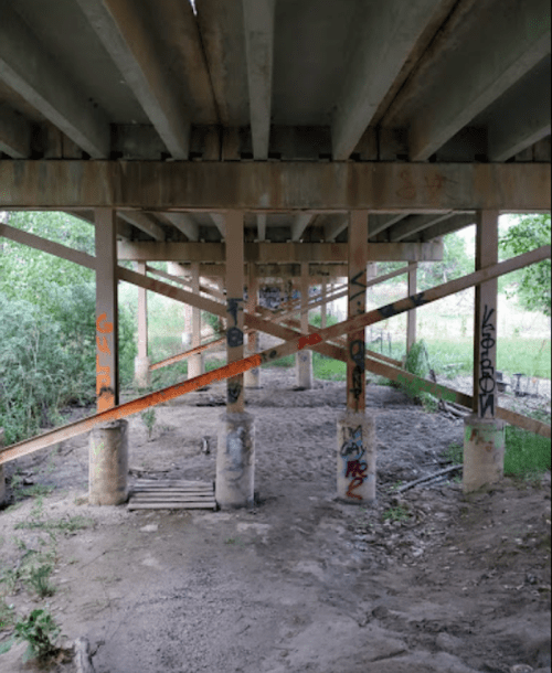 Under a bridge, concrete pillars are supported by wooden beams, surrounded by greenery and graffiti on the columns.