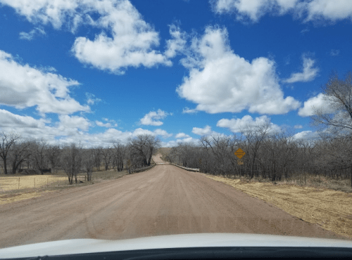 A dirt road stretches ahead under a blue sky with fluffy clouds, flanked by bare trees and warning signs.