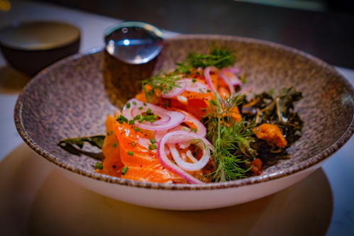 A bowl of salmon garnished with onions and herbs, served on a textured plate with a spoon beside it.
